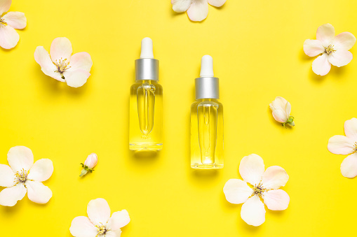 Spring concept of natural organic cosmetics, beauty herbal product spa aroma oil. Clean glass cosmetic bottles with dropper, delicate spring flowers on yellow background flat lay top view. Mockup.