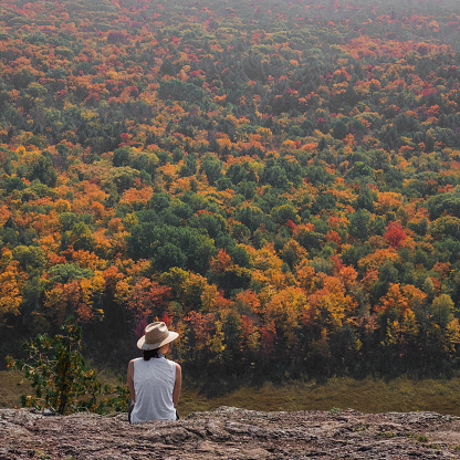 Bright Colorful Fall in Michigan UP, Lake of the Clouds, USA. A woman, wearing a straw hat and white shirt, is sitting on a rocky cliff, looking down at the bright, colorful fall trees.
