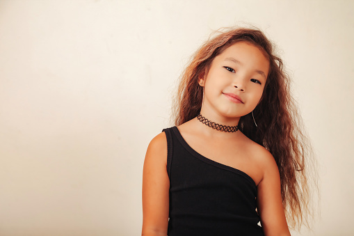 Lovely asian little girl with long curly hair posing at white background, looking at camera. Studio shot stylish funny small stylish model. Fashion education concept. Copy text space for advertising