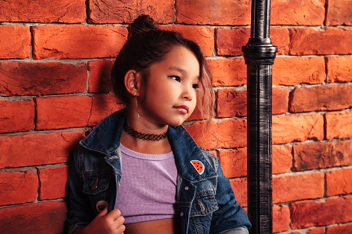 Portrait funny asian little girl in denim jacket posing at brick wall background, looking away. Small brunette model standing in district. Childhood style concept. Copy text space for advertising