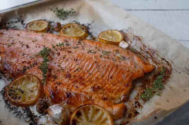 Delicious oven roasted or baked half salmon, mediterranean style. Cooked with thyme, garlic, olive oil and lemon. Served ready to eat on a baking sheet isolated on wooden background