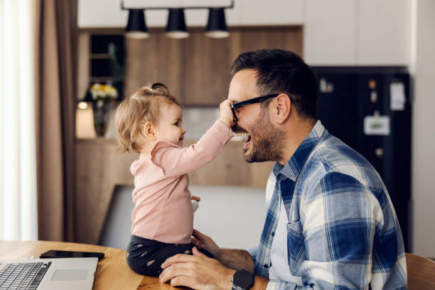 A silly baby girl is playing game with her dad. She is trying to take his eyeglasses on his work break. stock photo