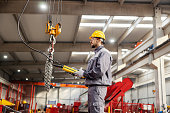 A heavy industry worker relocates chains on hook and commands while standing in a metalwork factory.