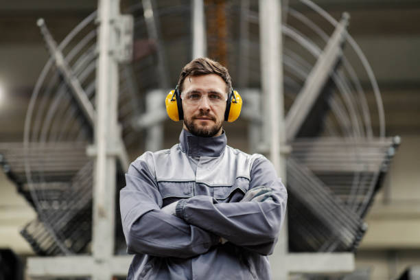istock A heavy industry worker with arms crossed makes eye contact while standing in a facility with big metal construction in the background. 1459018059