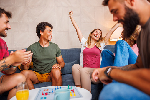 Group of cheerful young friends having fun playing ludo board game while spending time together at home