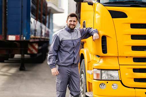 A lorry driver prepares for shipping abroad. He is leaning on a lorry and smiles at the camera.