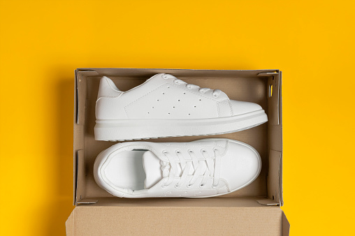White women's leather sneakers in cardboard box on yellow background top view flat lay. Stylish youth sneakers, sports shoes, genuine leather footwear. Minimalistic shoe store advertising, fashion.