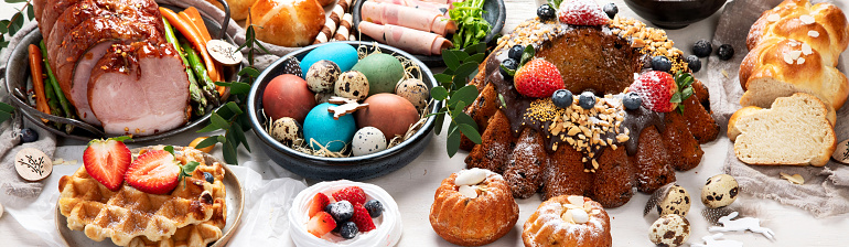 Traditional Easter dinner or  brunch with ham, colored eggs, hot cross buns, cake and vegetables. Easter meal dishes with holday decorations. Panorama, banner