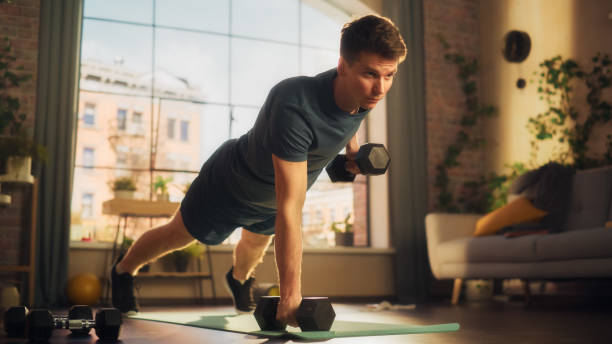 istock Fit Athletic Strong Young Man Performing Enduring Training in Plank Position, while Lifting Dumbbells During Morning Workout at Home in Cozy Bright Apartment. Concept of Healthy Lifestyle and Fitness. 1459025673
