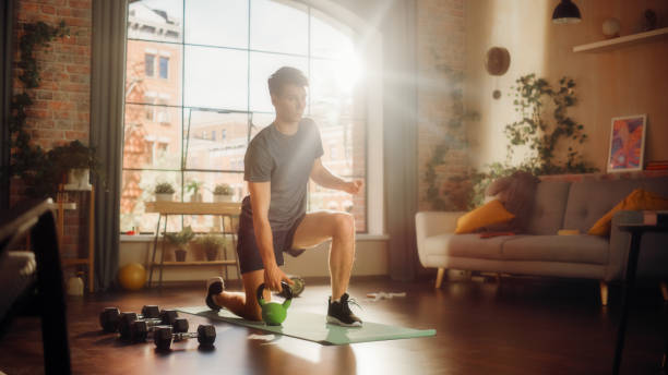 istock Strong Athletic Fit Young Man Standing on One Knee and Lifting a Heavy Kettlebell, Doing Core Strengthening Exercises During Morning Workout at Home in Sunny Apartment. Concept of Health and Fitness. 1459025794