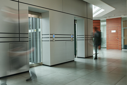 Part of corridor of modern office center with blurred silhouettes of two businessmen moving along two elevators with transparent doors