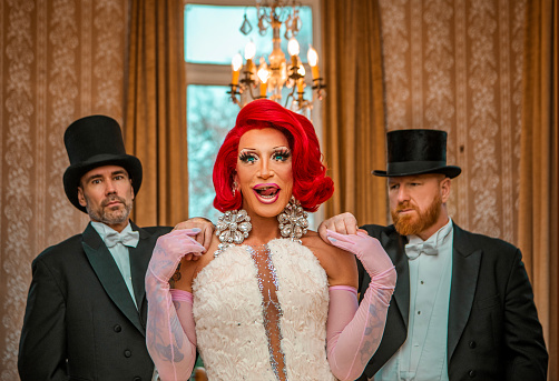 A beautiful elegant 1920s style redhead drag queen in a luxury stately home with handsome male chaperones