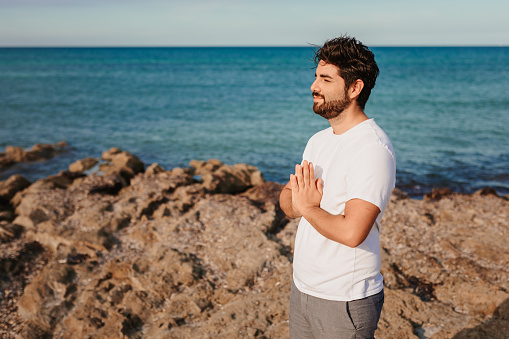 Praying handsome man with hands on chest and grateful face gesture in front of the sea. Useful for healthy, mindfulness, consciousness, awareness and self love concepts. Very selective focus and added grain. Part of a series.