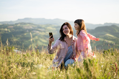 Mother with daughter taking selfie when sitting on grassy hillside with Mountain View in italy. Happy hiker girl resting on the grass in the mountains and taking photos.
