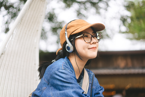 People mental therapy with at outdoor nature lifestyle concept. Young asian woman relax listening music with fashion headphone wearing hat denim jacket and eyeglasses.