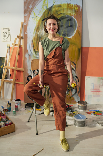 Young smiling female artist or painter in jumpsuit sitting in the center of home studio against panel with masterpiece and looking at camera
