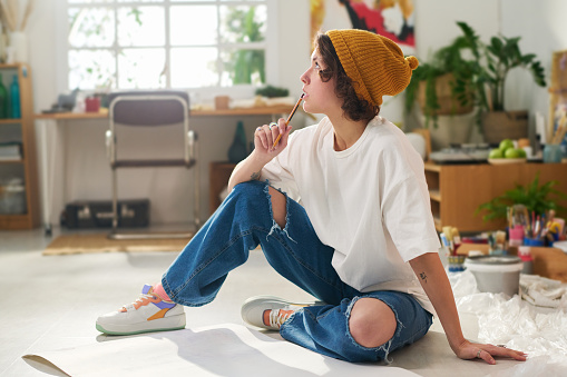 Young pensive woman in casualwear holding pencil by her mouth while sitting on the floor with papers and thinking of new artwork