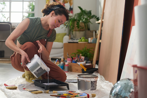 Young creative female artist pouring liquid paint into plastic tray before working over creation of new masterpiece in home studio