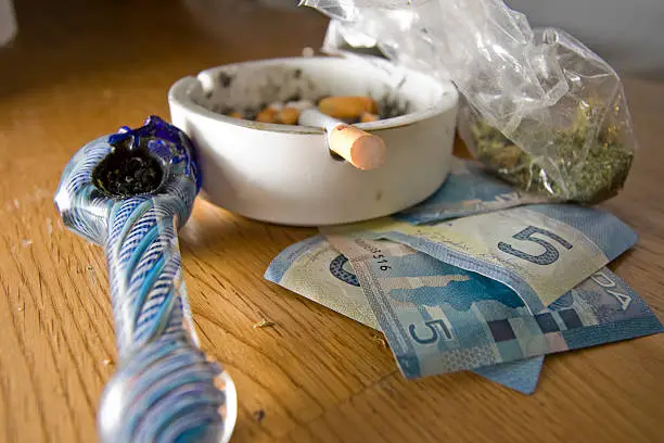 conceptual photo of a small pile of cash with a pipe, ashtray with cigarettes and small bag of herbal narcotics.