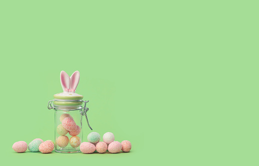 Easter bunny ears sweets jar with many tiny eggs in and near on green edgeless background. Happy Easter holiday concept. Easter chocolate treats web banner