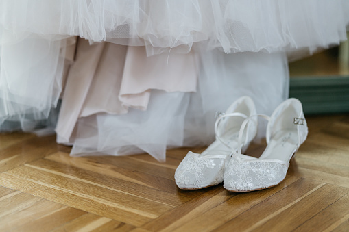 white wedding shoes of bride standing on wooden floor in front of wedding dress