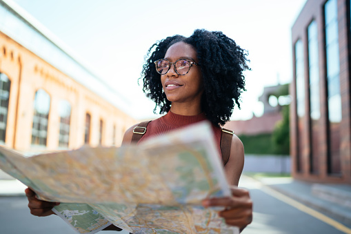 Young black lady wearing glasses and backpack smiling and studying map while looking away and walking on territory outside of campus