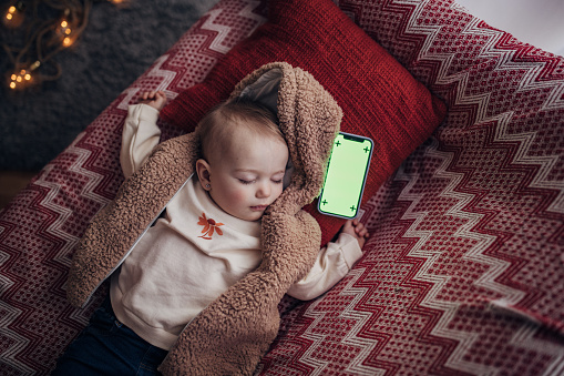 Beautiful baby girl is sleeping on sofa in her home, smart phone is next to her head.