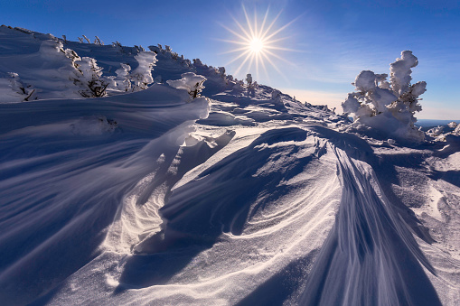 The sun shines above the snowy summit of Algonquin Peak, in the  Adirondack High Peaks of New York State, on a bluebird winter day. The high winds that the alpine summit is exposed to carved the snow into interesting shapes and patterns and coated the alpine trees with rime ice. A small aperture was used to render the sun as a sunstar or sunburst. Algonquin Peak is one of the Adirondack 46 High Peaks, which are mountains above 4,000 feet in elevation. Climbing all 46 peaks and becoming a 46er is a popular hiking challenge.