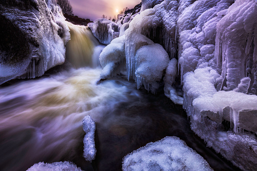 Cold winter temperatures caused the spray from a waterfall in the Adirondacks to freeze and form beautiful intricate ice formations along the edge of the waterfall. The sun is beginning to set in the background, and the presence of tannins in the water can be seen by the tan and brown tint to the water.