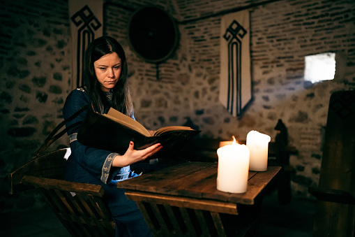 Medieval woman sitting by the desk, and reading book with a help of candlelight