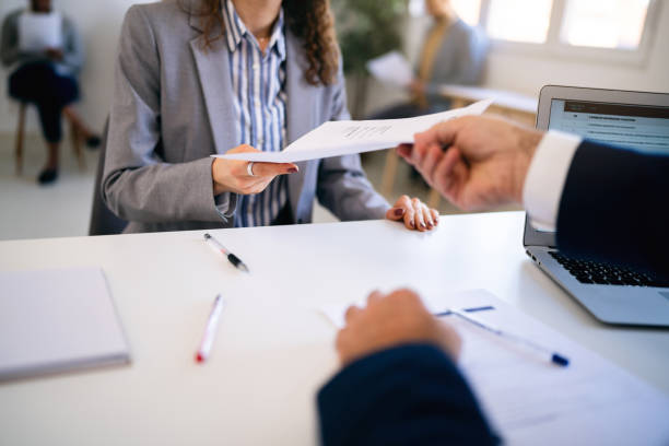 Female candidate, handing to a recruiter her resume during job interview In the office young Caucasian female candidate, giving her CV or application form to male recruiter during a job interview 
Other candidates sit on the chair and wait for their turn employment document stock pictures, royalty-free photos & images