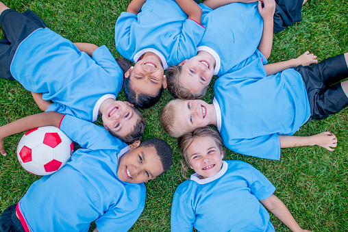 A small group of 6 school aged children lay in a circle n the ground with their heads together, as they pose for a portrait.  They are each wearing light blue jerseys and smiling in their aerial photo.