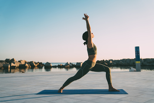 Side view of slim young woman with brown hair in sportswear making crescent lunge pose on fitness mat against background of sky