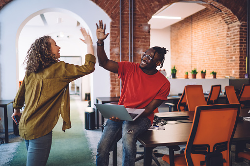 Cheerful young diverse colleagues celebrating successfully completed project and giving high five while working together in modern loft conference room