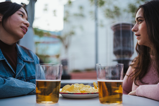 Two young women having a discussion at a bar terrace while having a snack and beer during a break