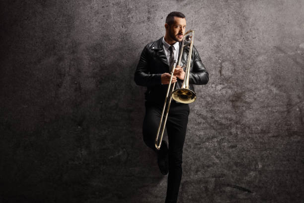 istock Man in a leather jacket holding a trombone and standing against a gray wall 1459116043