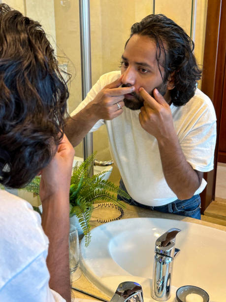 istock Image reflection in mirror of Indian man stood at sink in bathroom looking at himself squeezing a spot, skincare pimple popping, artificial fern plant in metal pot on marble bathroom counter, bathroom sink, toothbrushes in glass 1459116883