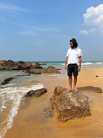 Stock photo showing beach coastal rock feature at golden sandy beach with man standing at the top of boulder looking at sea.