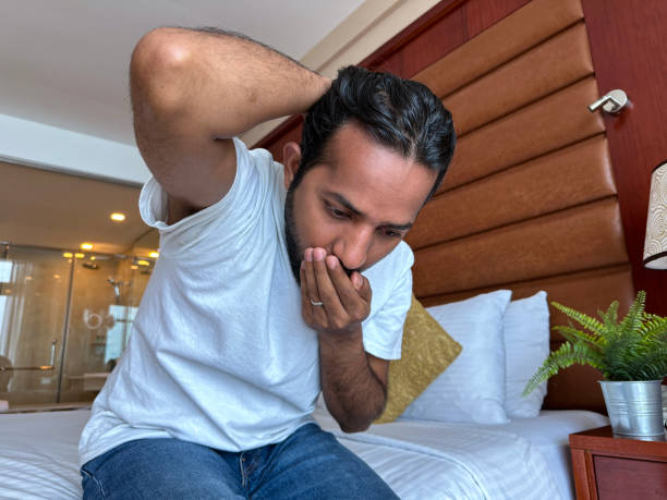 imagem em close-up do homem indiano sentado ao lado da cama de casal do hotel segurando a mão sobre a boca para parar de estar doente, segurando o cabelo para evitar o vômito, criado-mudo com vaso de planta e lâmpada, foco em primeiro plano, feriado e c - double bed night table headboard bed - fotografias e filmes do acervo