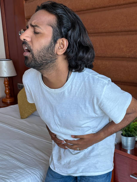istock Close-up image of Indian man standing besides hotel double bed holding both hands over lower stomach, doubling over in pain, night stand with potted plant, focus on foreground, holiday and vacation illness concept 1459126739