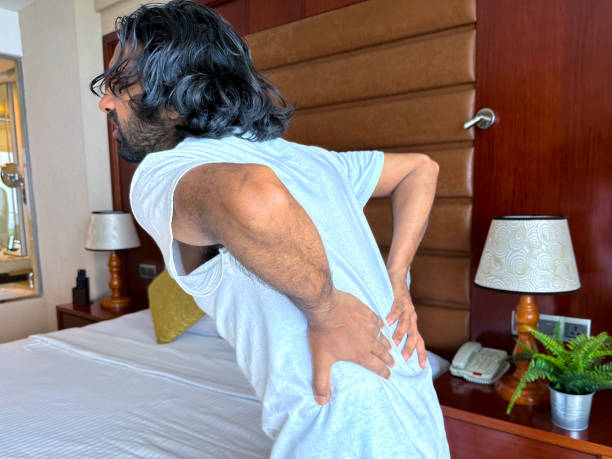 istock Close-up image of Indian man standing besides hotel double bed holding both hands over lower back, doubling over in pain, backache, night stand with potted plant, telephone, lamp focus on foreground, holiday and vacation illness concept 1459127342