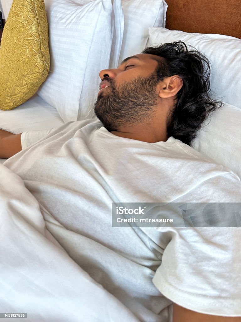 Close-up image of Indian man lying on pillow in hotel room double bed, snoring and sleeping under white bed sheet, leatherette headboard, focus on foreground Stock photo showing close-up view of man lying under bedcovers in double bed snoring whilst sleeping. 30-34 Years Stock Photo Close-up image of Indian man lying on pillow in hotel room double bed, snoring and sleeping under white bed sheet, leatherette headboard, focus on foreground Stock photo showing close-up view of man lying under bedcovers in double bed snoring whilst sleeping. 30-34 Years Stock Photo