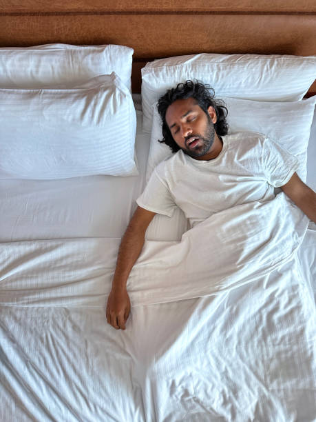Image of Indian man lying on pillow in hotel room double bed, snoring and sleeping under white bed sheet, leatherette headboard, elevated view, copy space stock photo