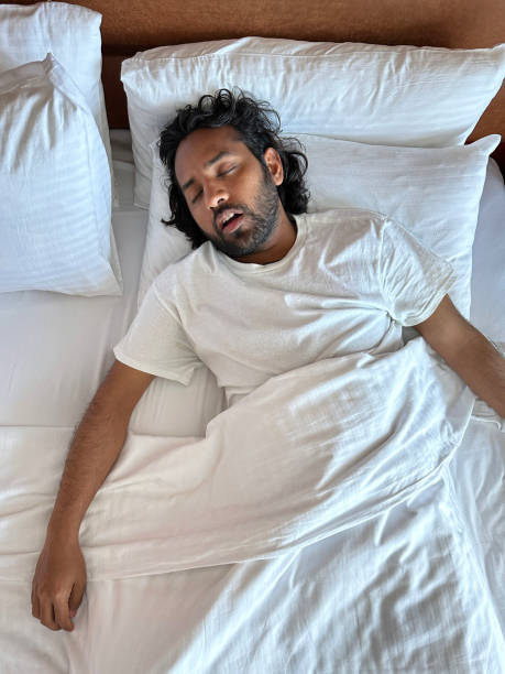 Close-up image of Indian man lying on pillow in hotel room double bed, snoring and sleeping under white bed sheet, leatherette headboard, elevated view Stock photo showing close-up, elevated view of man lying under bedcovers in double bed snoring whilst sleeping. man sleeping top view stock pictures, royalty-free photos & images