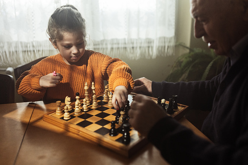 Old man playing chess with his little granddaughter in living room at home.