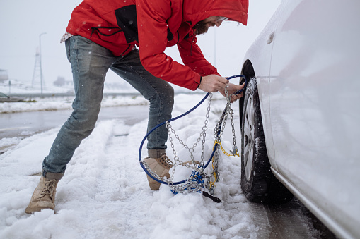 A low angle view of a front wheel drive vehicle on a snowy and ice covered road, a man putting chains on the wheels to improve traction in the winter weather conditions. Horizontal image with copy space.