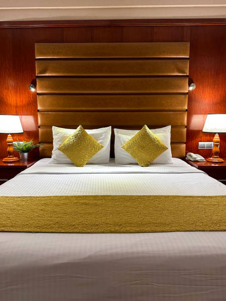 imagem da cama de casal do quarto de hotel com pilha de travesseiros brancos do hotel apoiados na cabeceira de couro sintético, capa de edredom branco, almofadas de ouro, lâmpadas elétricas iluminadas em mesas de cabeceira, foco em primeiro plano - double bed night table headboard bed - fotografias e filmes do acervo