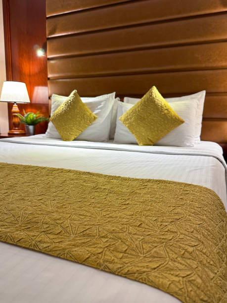 istock Close-up image of hotel room double bed with stack of white hotel pillows leaning on leatherette headboard, white duvet cover, gold cushions, illuminated electric lamp on night stand, focus on foreground 1459137591