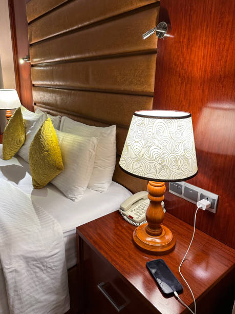 imagem em close-up de lâmpada elétrica iluminada, telefone e smartphone de carregamento em criado-mudo, além de cama de casal do hotel com capa de edredom branco, pilha de travesseiros brancos do hotel apoiados na cabeceira de couro sintético, almofada - double bed night table headboard bed - fotografias e filmes do acervo