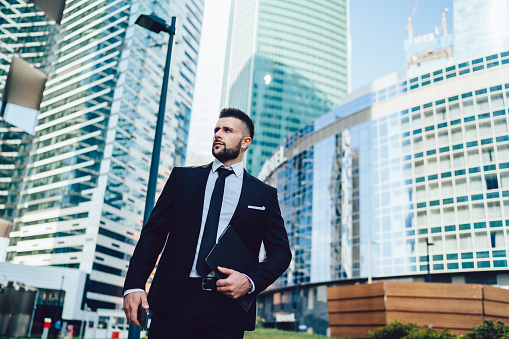 From below view of assured thoughtful young businessman with folder and smartphone walking in megapolis centre against modern high buildings looking away